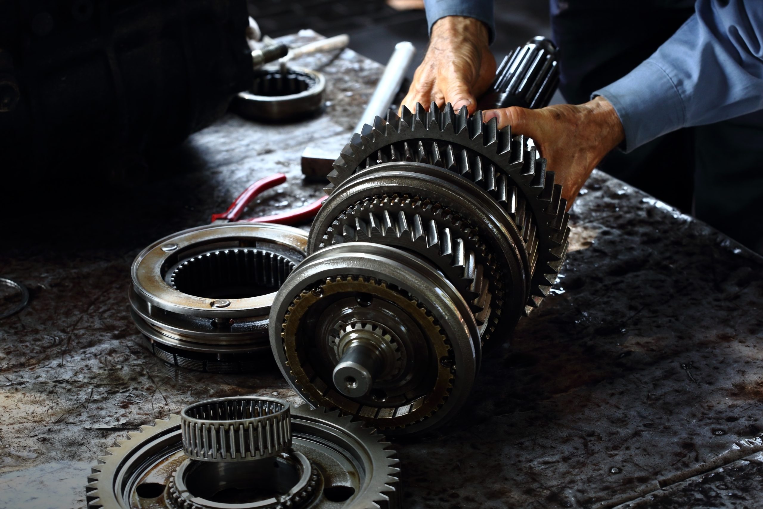 Mechanic assembling a complex vehicle transmission gear shaft on a cluttered, oily workbench.